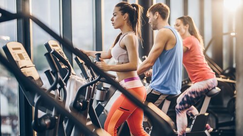 Das Bild zeigt ein Fitnessstudio von innen und 3 Menschen, die am Laufband stehen.
